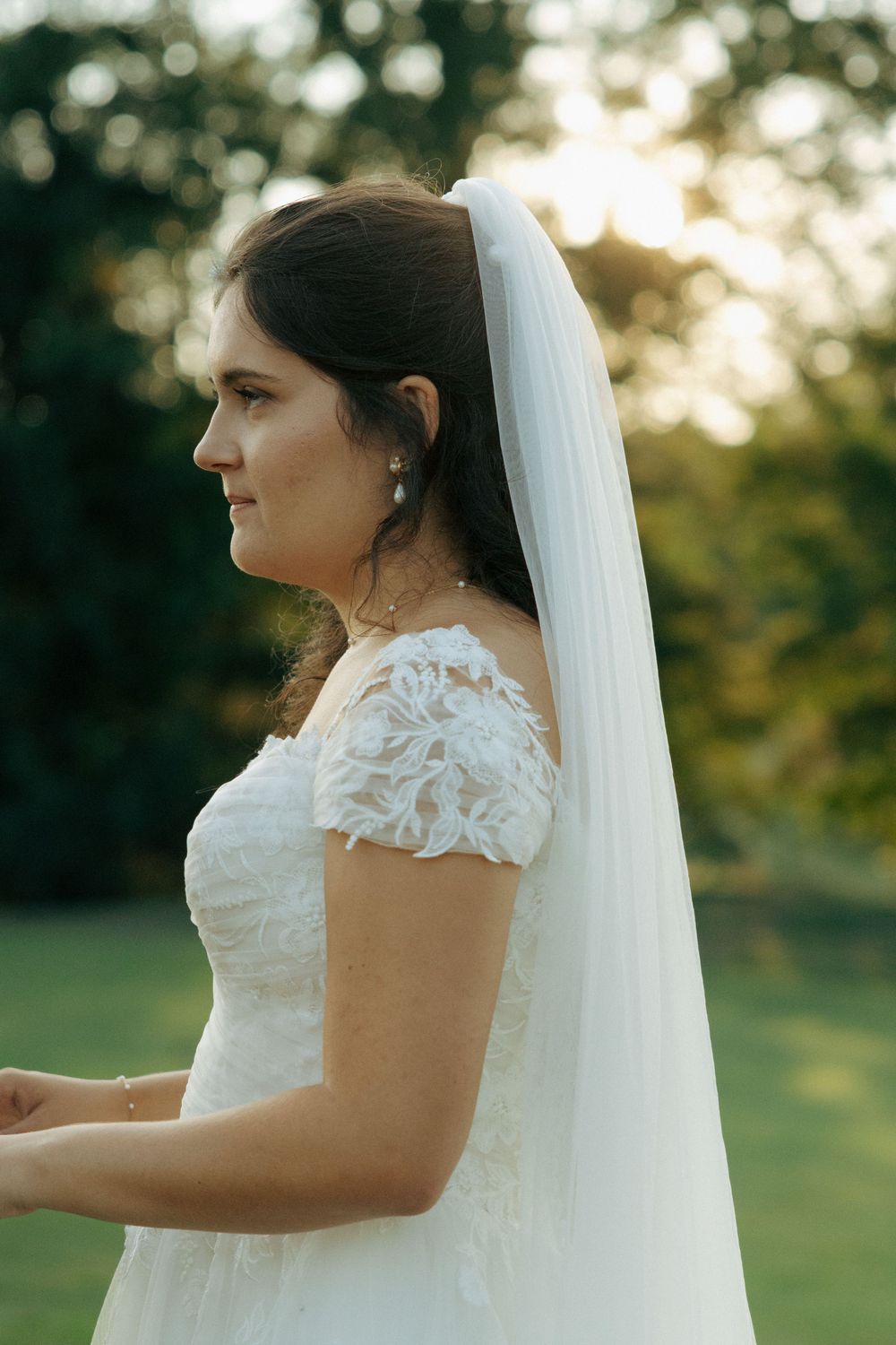 A bride in a white lace dress with cap sleeves and veil poses against a sunset-lit outdoor background.
