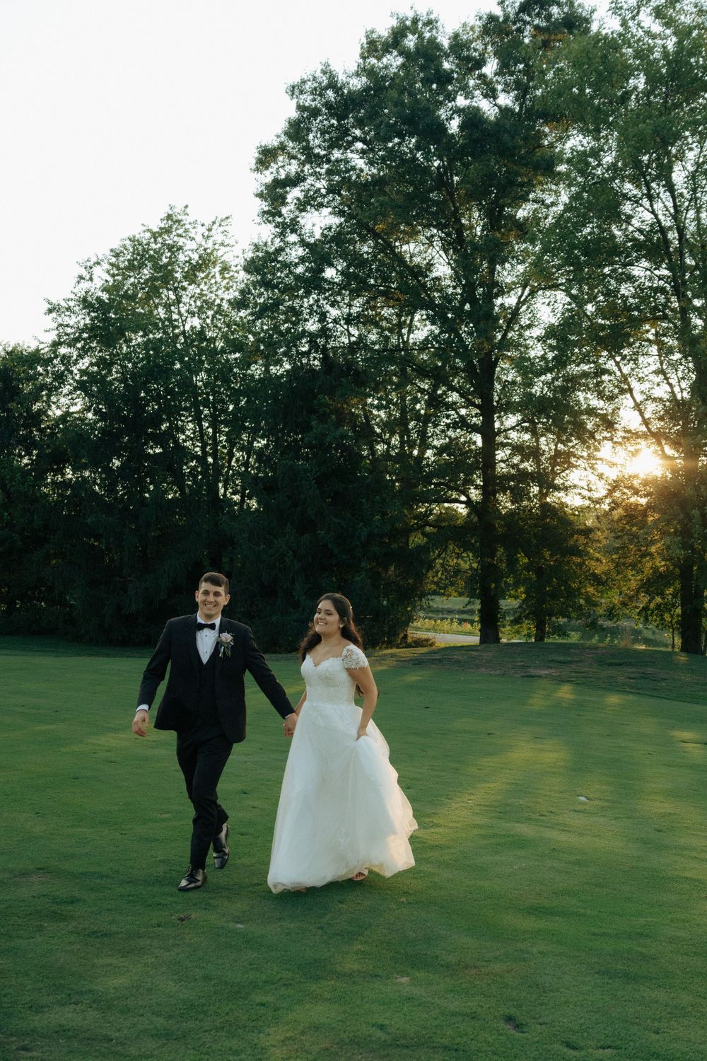A newlywed couple runs playfully across a green lawn with trees and golden sunset light in the background.