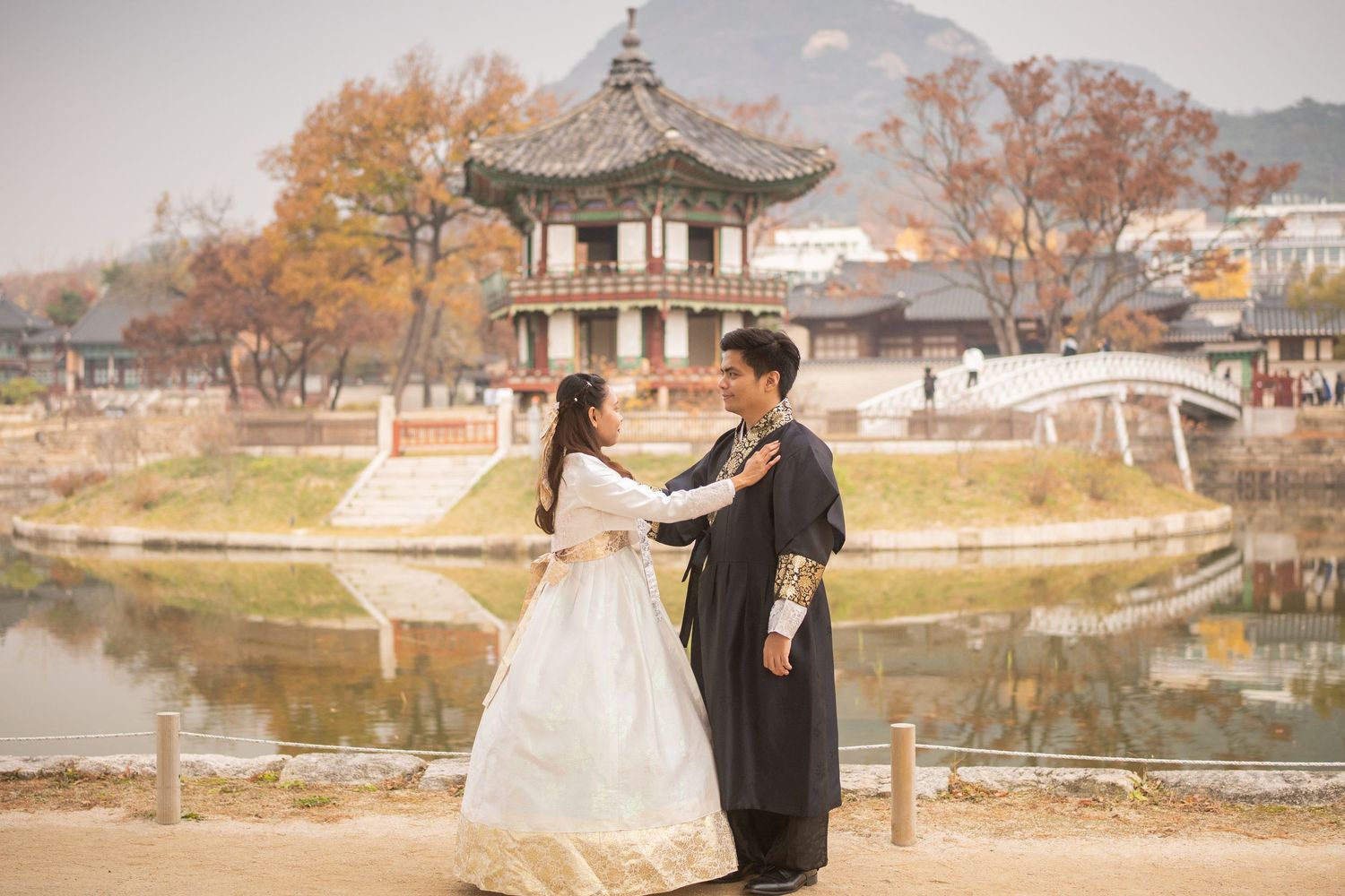 Couple in wedding attire poses near a tranquil lake with traditional Korean architecture.