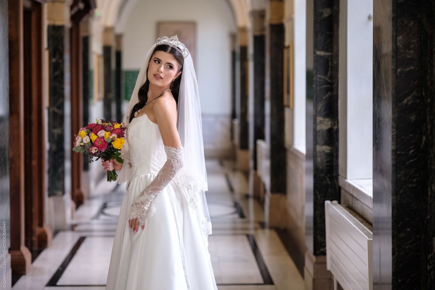 Bride in elegant white lace dress poses in grand hallway with arched doorways and natural lighting.