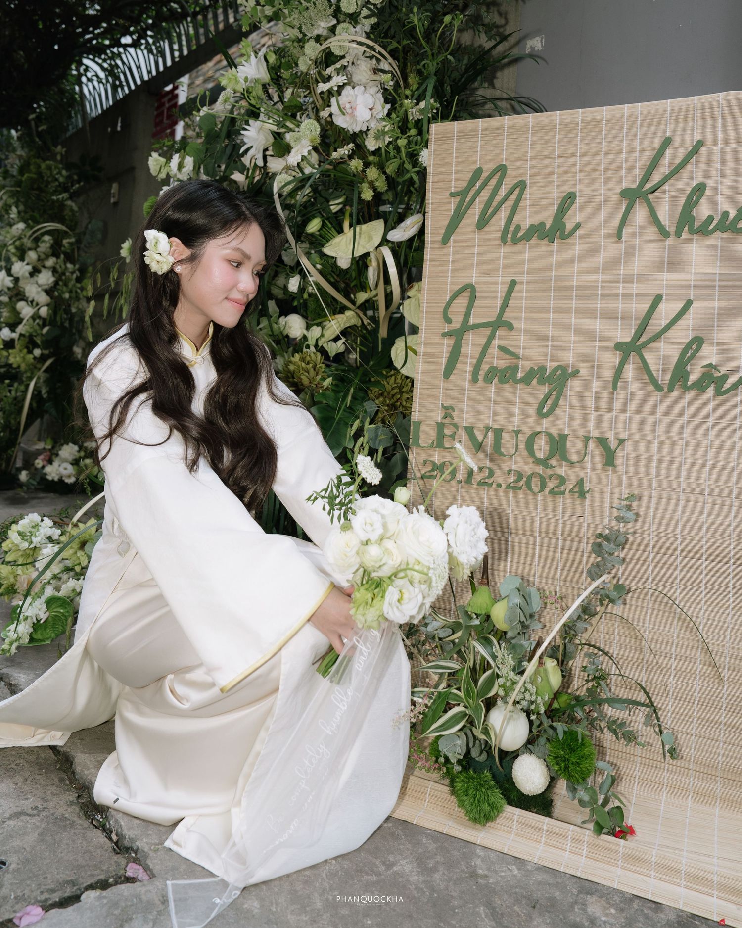 Vietnamese bride in white ao dai kneels beside bamboo wedding sign surrounded by white flowers.
