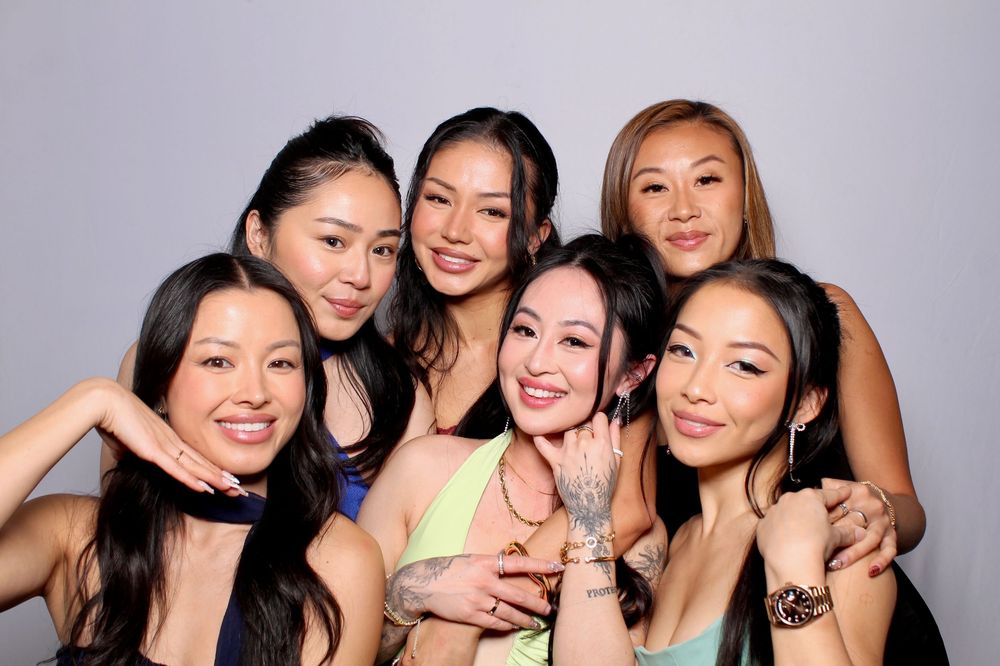 A group of five women pose together smiling in a photo booth setting with a gray backdrop.