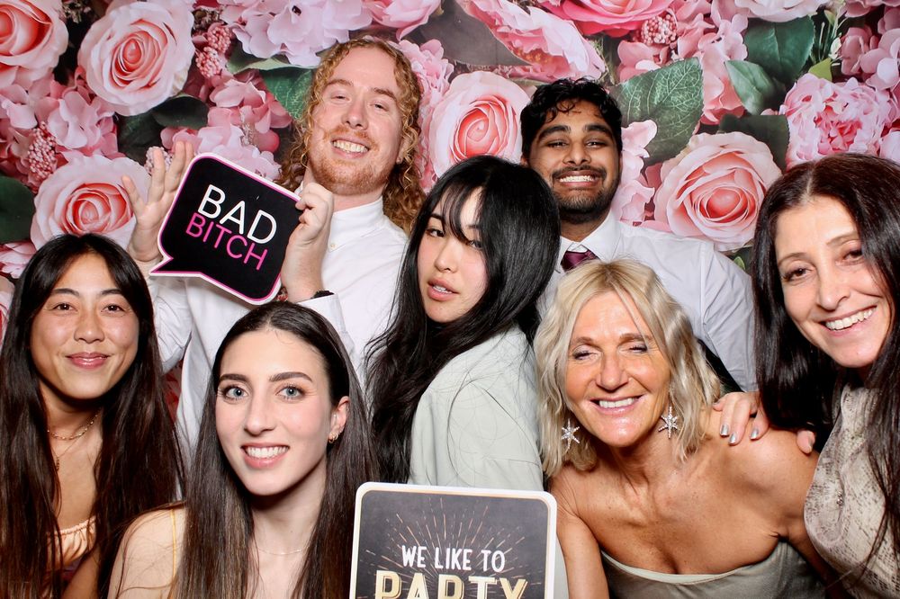 Group of happy people posing together at a party with pink floral decor and signs in the background.