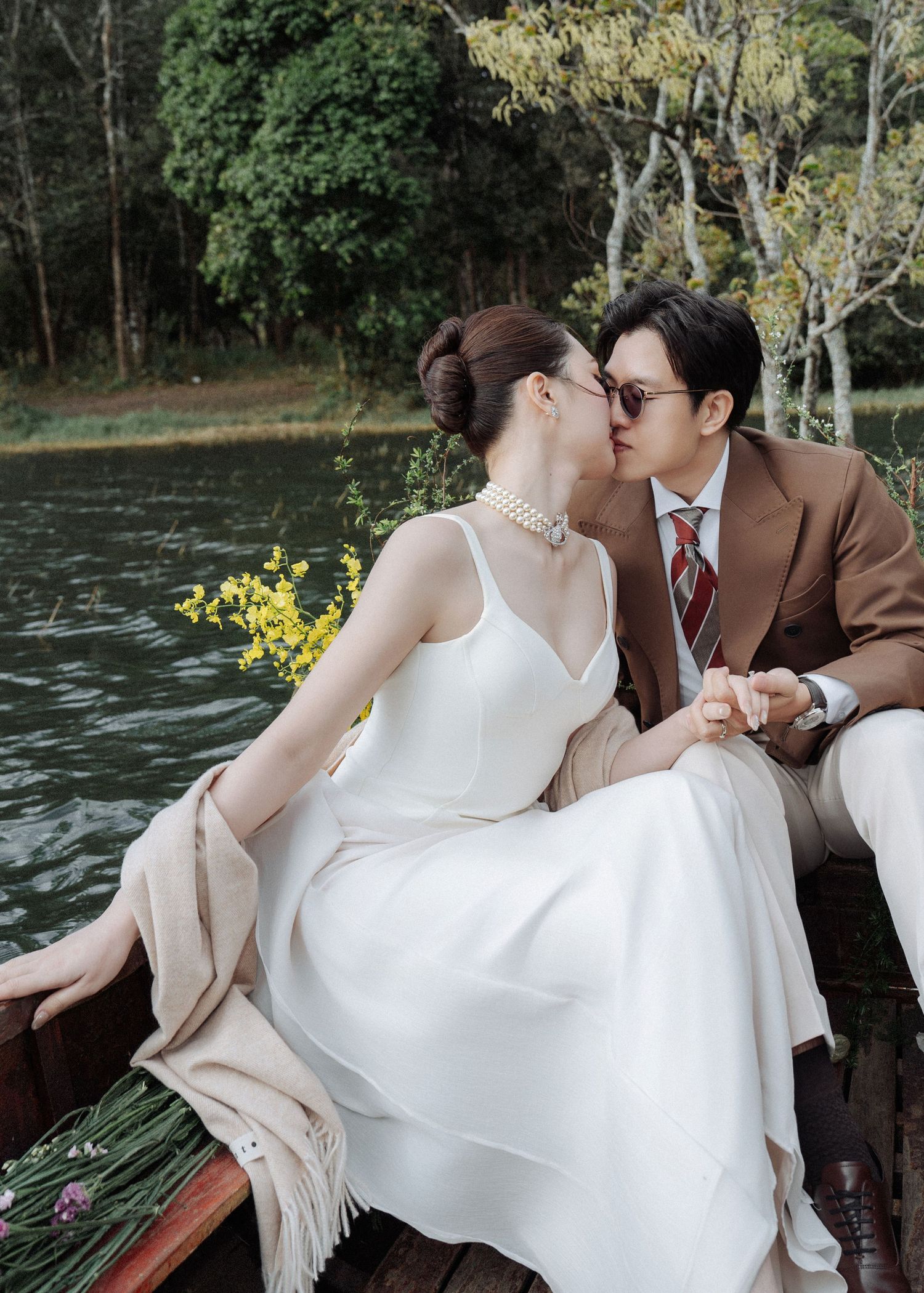Bride and groom share a kiss in flower-adorned wooden boat on tranquil lake surrounded by lush forest.