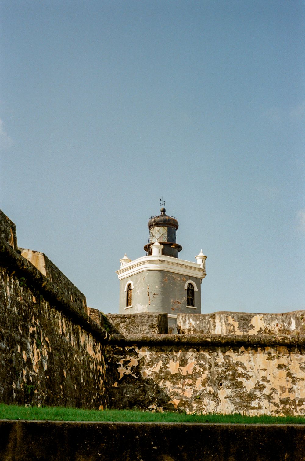 A series of weathered stone fortress walls with a white lighthouse tower and blue sky in the background.