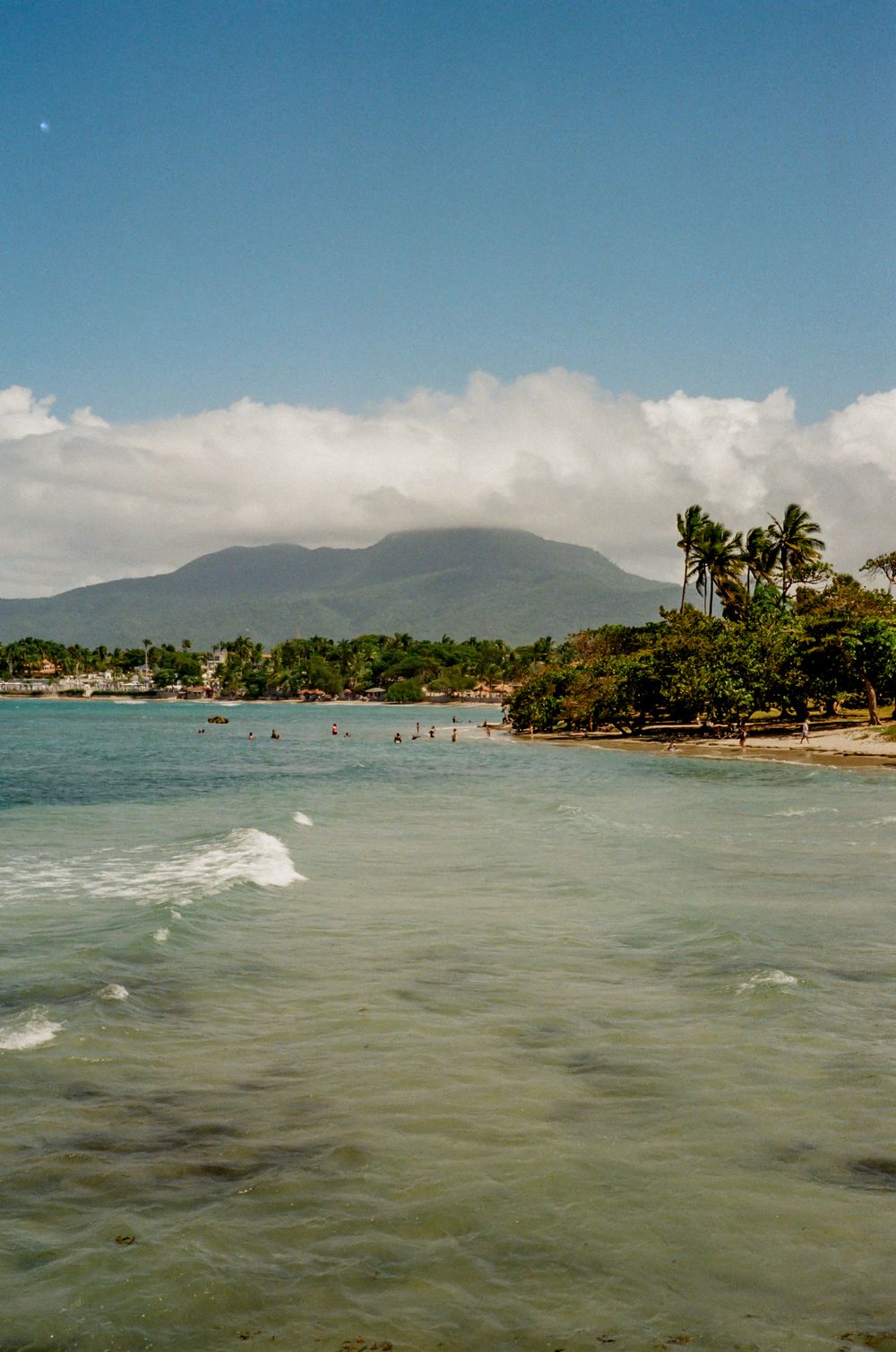 Panoramic view of a tropical beach shoreline with palm trees and mountains in the background under blue skies.