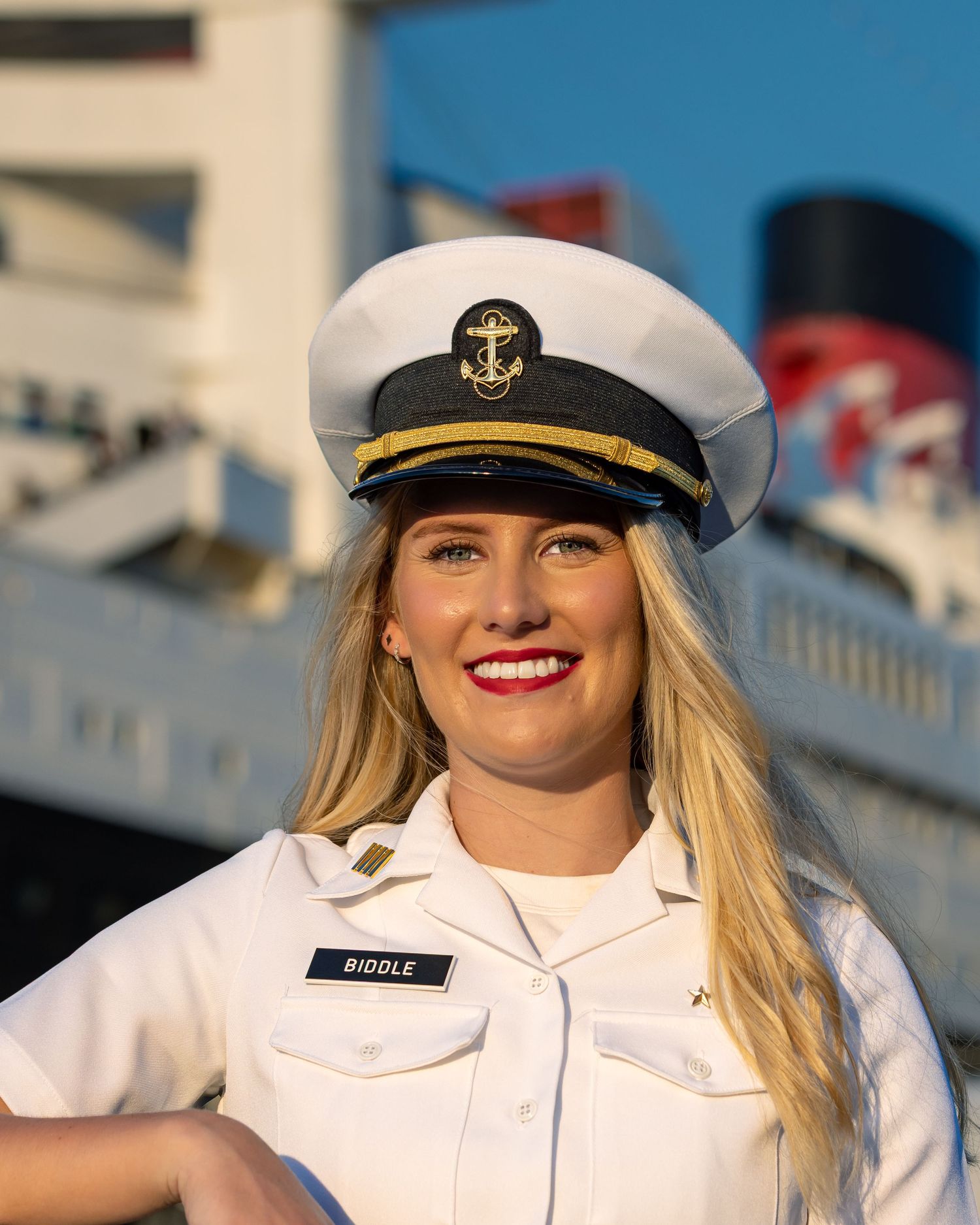 A person in a white naval uniform and captain's hat smiles while standing outdoors.