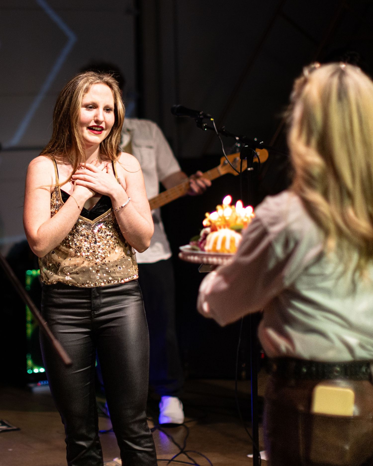 A daughter in a leopard print top and black pants gestures expressively as her mother brings her birthday cake.