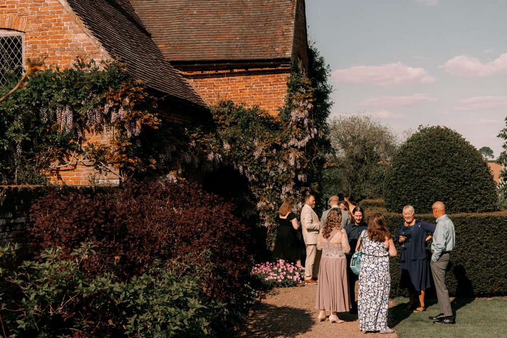 A group of wedding guests gather outside a rustic brick building with ivy and manicured hedges in the garden.
