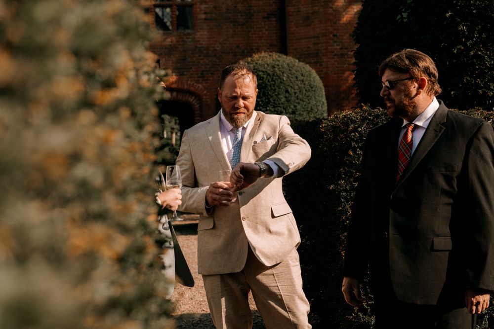 Two men in formal suits stand together near greenery and a brick wall at an outdoor event.