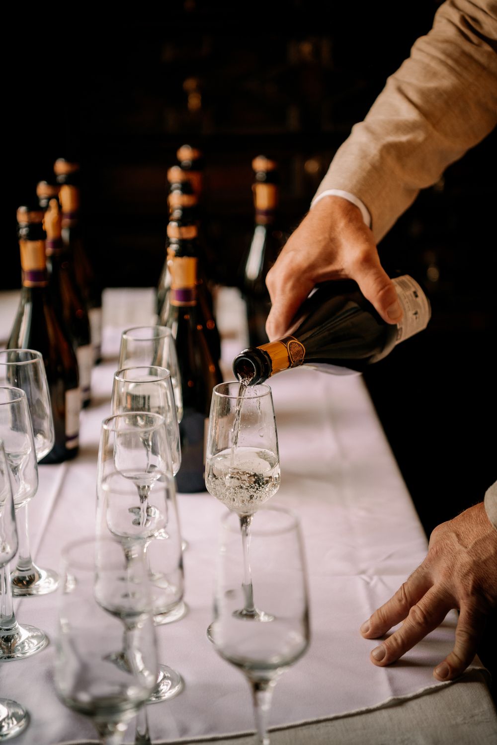Hands arranging empty wine glasses in a line on a white tablecloth in a dimly lit setting.