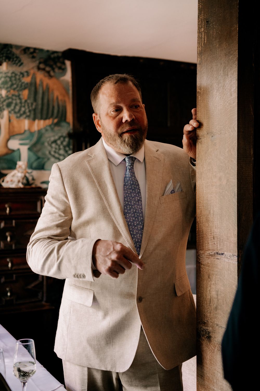 A person wearing a cream-colored suit and polka dot tie stands beside a wooden pillar in an elegant indoor setting.