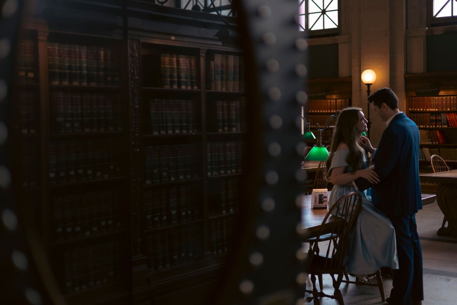 A couple studies together in a dimly lit vintage library with green desk lamp and wooden bookshelves in the background.