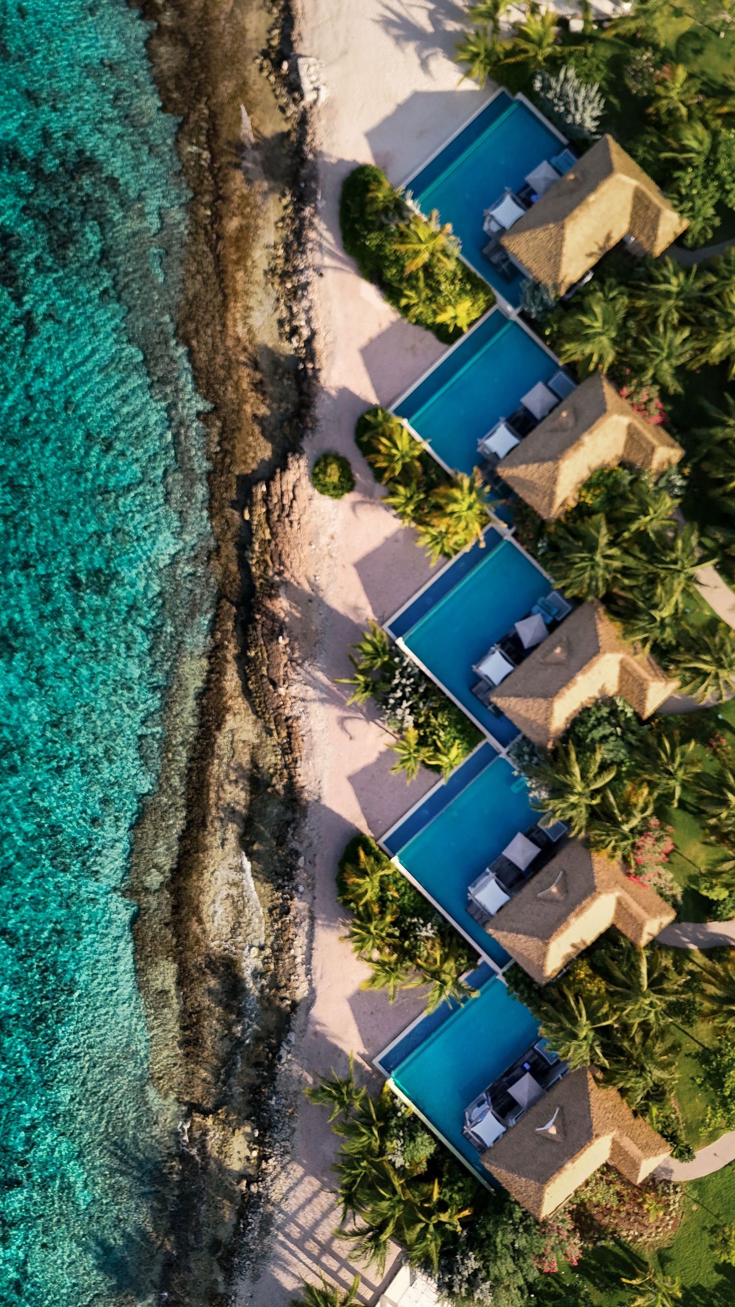 Aerial view of luxury beachfront villas with private pools along turquoise waters.