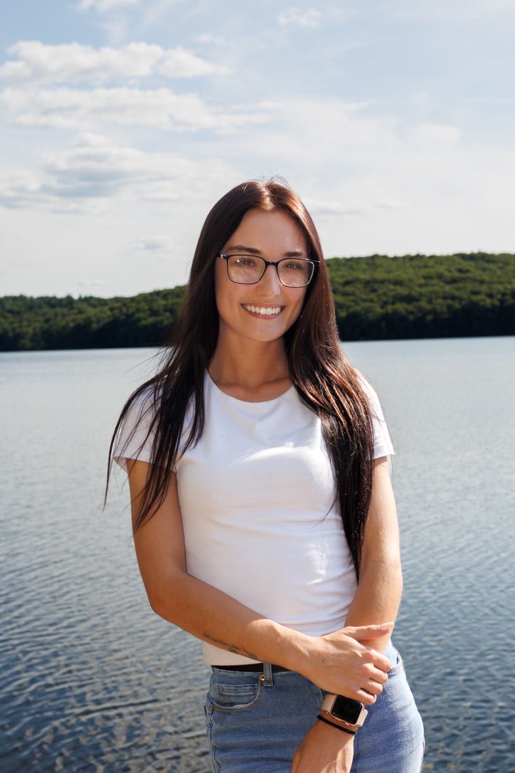 A woman in a white t-shirt and jeans poses by a scenic lake with mountains and forest in the background.
