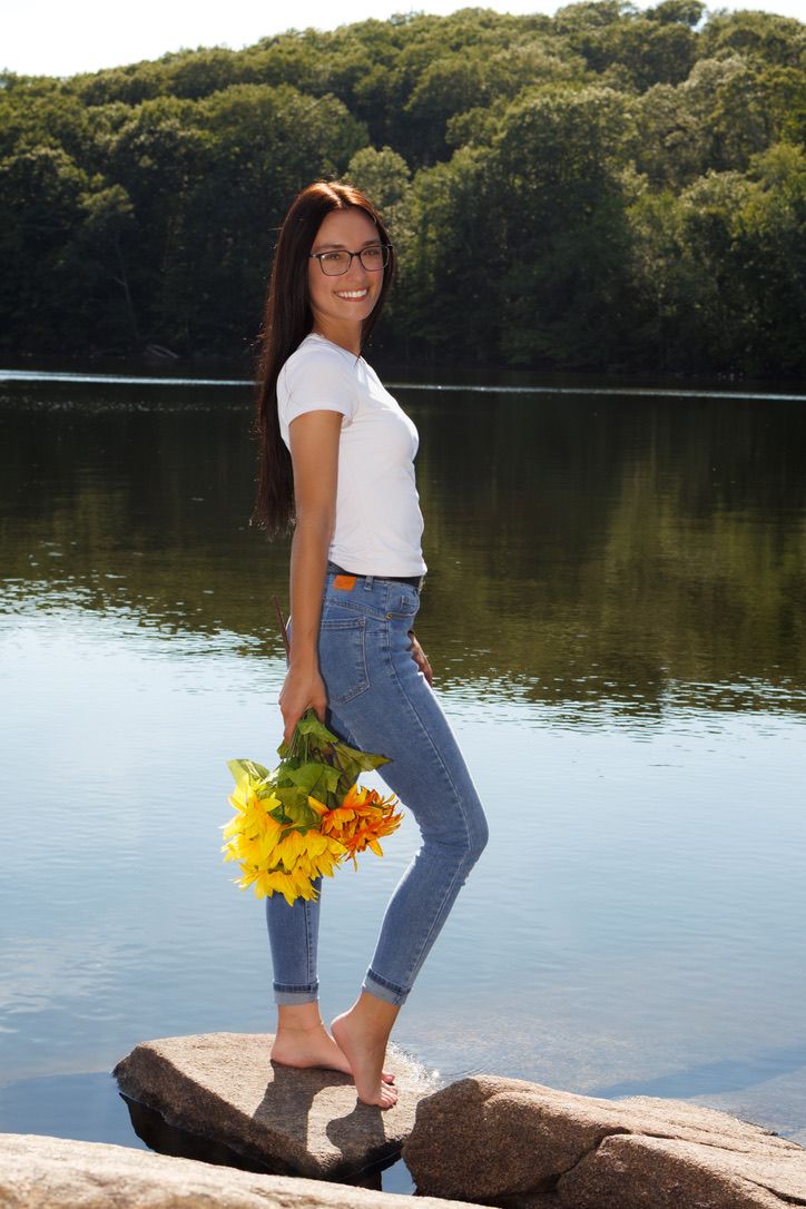 A person in jeans and white top holds yellow sunflowers while standing near a tranquil lake on rocks at sunset.