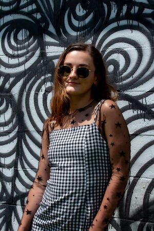 A person in sunglasses and a gingham tank top poses against a black and white swirl patterned wall.