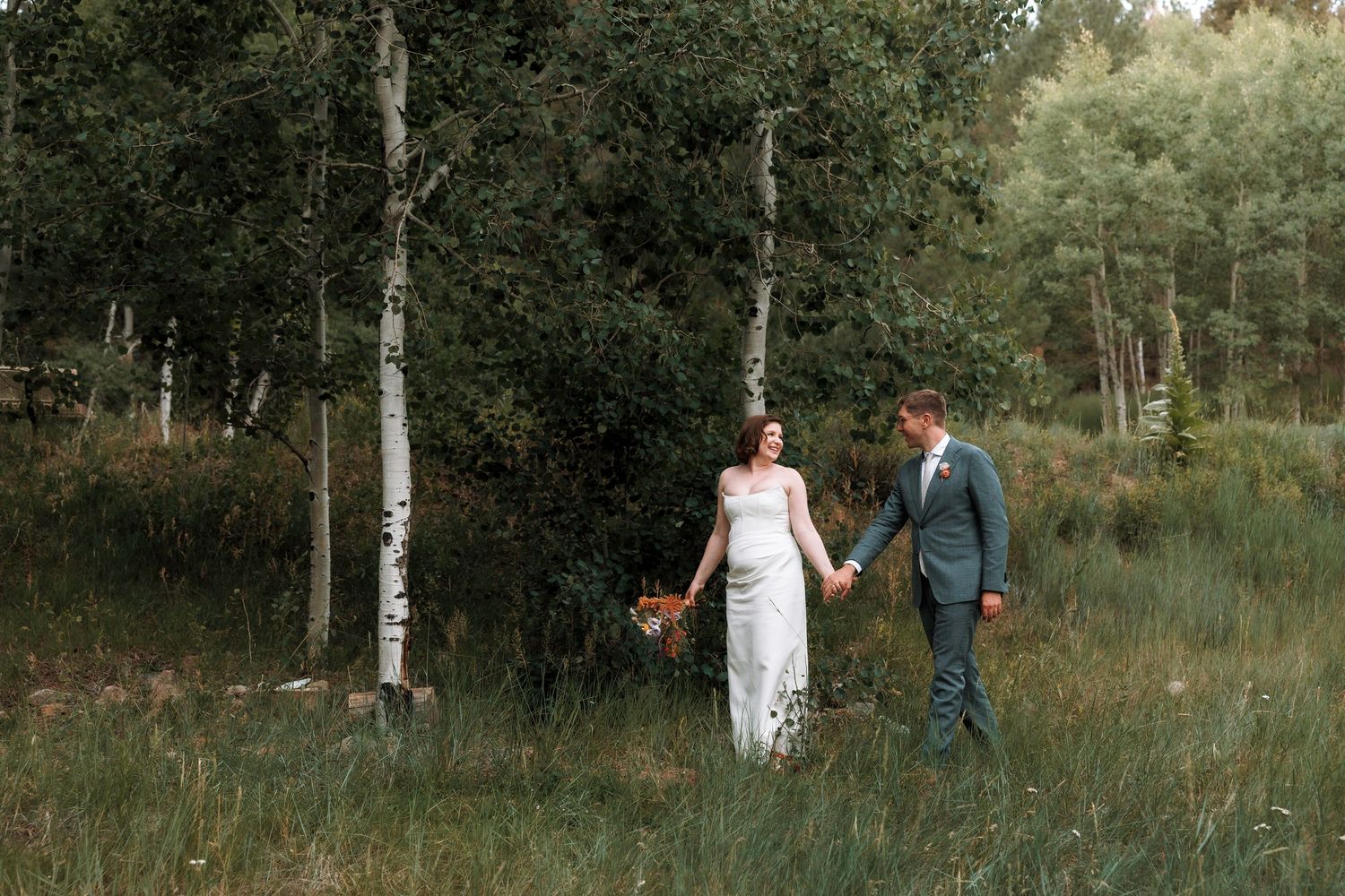 Bride and groom holding hands walking through tall grass among birch trees in forest.