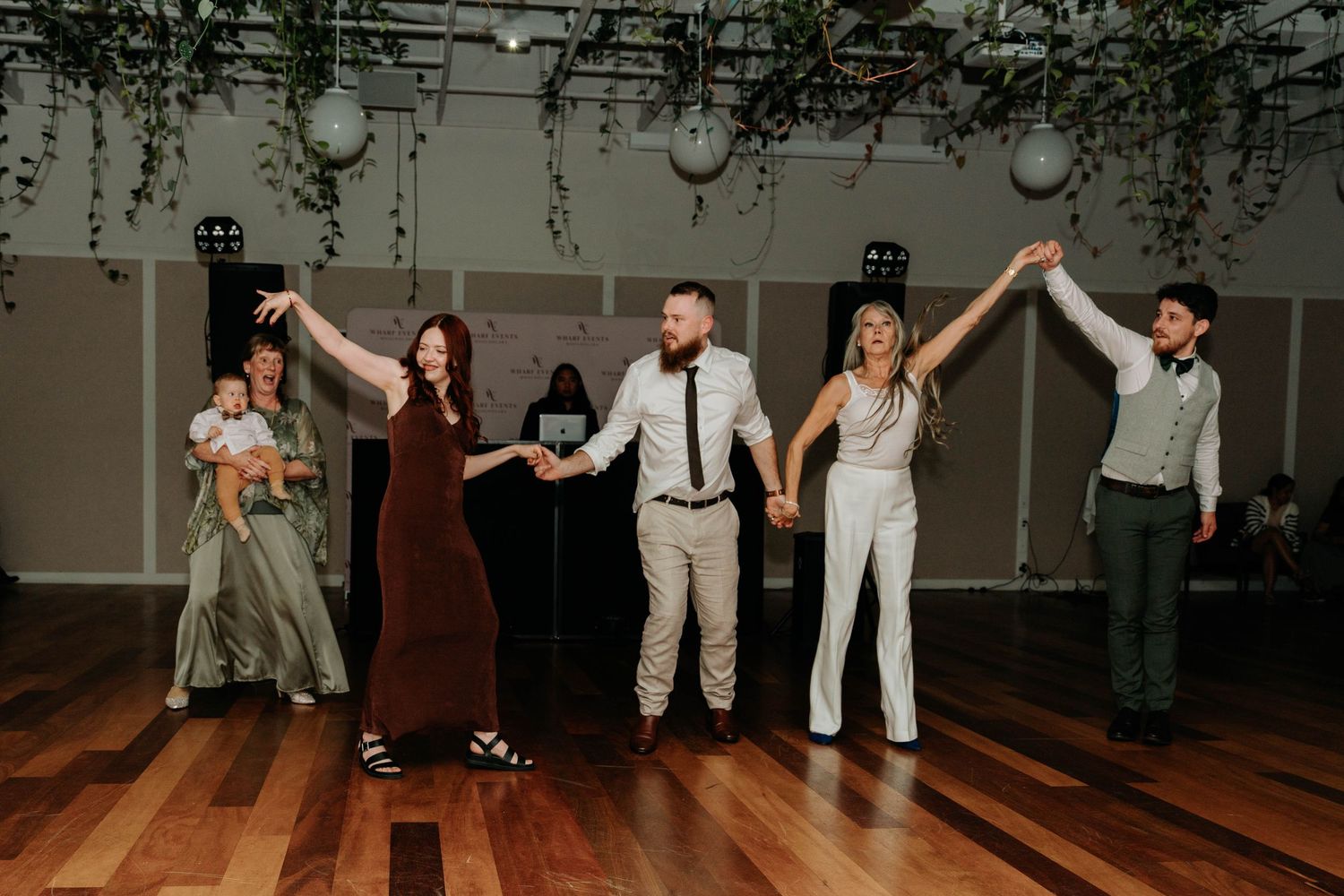A group of people dancing and holding hands in a line on a wooden dance floor under hanging decorative lights.
