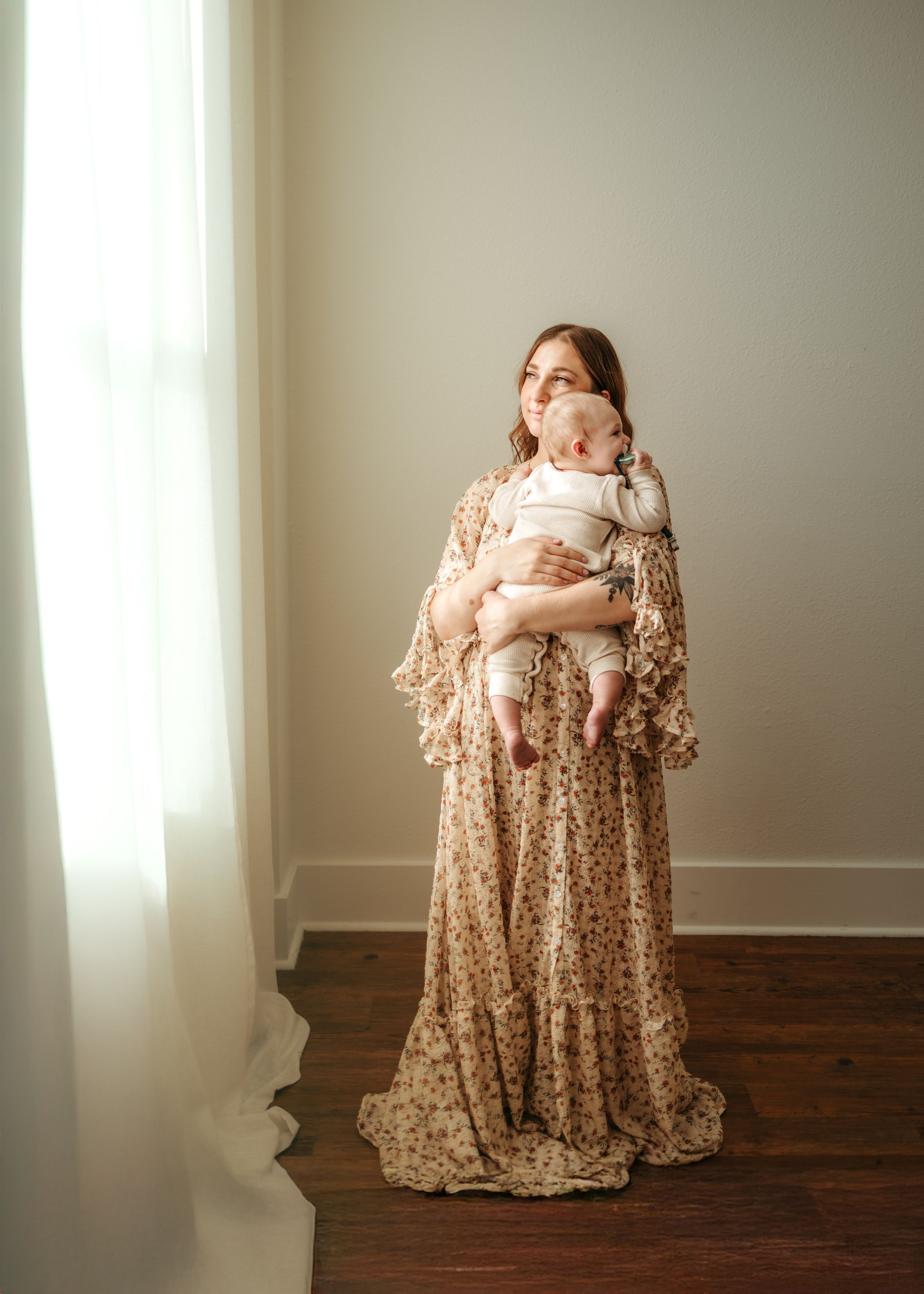 A newborn baby swaddled in white is cradled in a woman's arms wearing a flowing floral dress against a neutral backdrop.