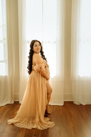 A person in a flowing peach-colored dress poses gracefully in front of sheer curtains on a hardwood floor.