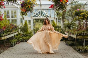 A person in a flowing peach gown poses in a greenhouse filled with vibrant hanging flowers and greenery.