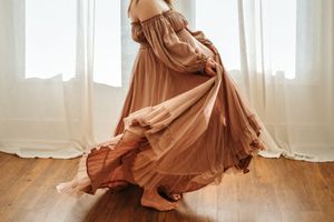 A flowing brown dress twirls gracefully in a sunlit room with white curtains and wooden floors.