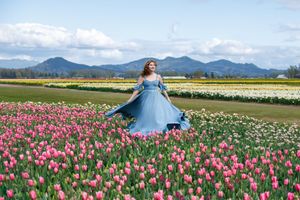 A person in a flowing blue dress twirls among vibrant pink tulip fields with mountains in the background.