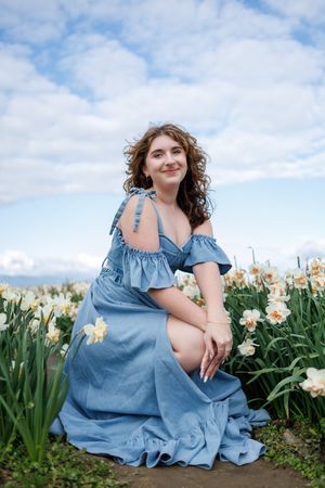 A person in a blue off-shoulder dress poses in a field of blooming daffodils under a cloudy spring sky.