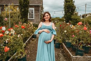 A woman in a turquoise dress stands in a blooming rose garden with red flowers and greenery surrounding her.