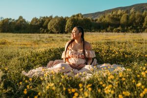 Someone in a floral dress lies in a golden wildflower meadow during sunset with mountains in the background.