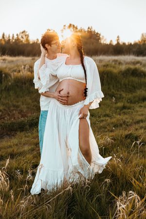 Maternity photo of a couple embracing in a golden sunlit field at sunset wearing flowing white outfits.