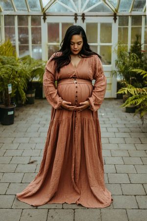 Expectant mother in flowing rust-colored maxi dress poses in front of greenhouse with potted ferns.