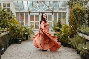 A flowing terracotta dress twirls elegantly in a sunlit greenhouse filled with lush plants and greenery.