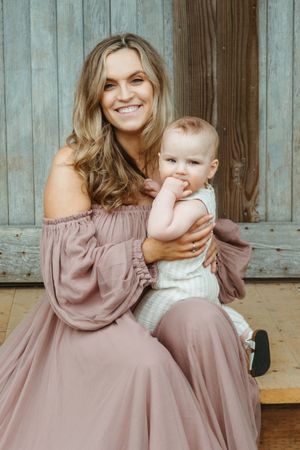 A parent in a mauve off-shoulder dress sits with a baby in white against a rustic wooden backdrop.