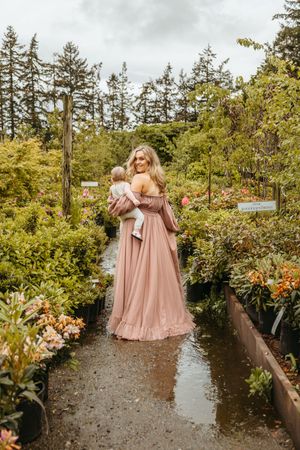 Bridesmaids in flowing blush pink dresses walk along a garden path surrounded by lush greenery and trees.