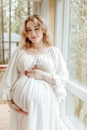 A woman in a flowing white maternity dress stands by large windows in a sunlit room with natural surroundings.