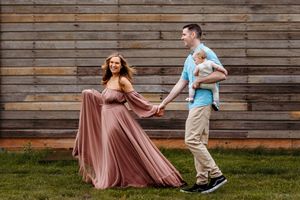 A couple dances gracefully on grass against a rustic wooden wall backdrop, her flowing mauve dress swirling with movement.