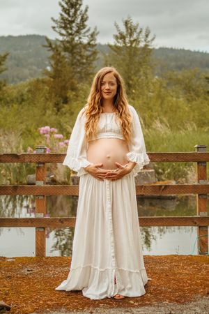 A maternity photoshoot in a white lace dress shows a pregnant woman standing by a wooden fence near a lake.