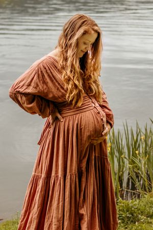 Person in flowing rust-colored tiered dress stands near water's edge with tall grass and reflective lake surface.