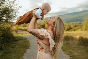 Parent in beige dress holds and snuggles with baby against scenic mountain landscape backdrop during sunset.