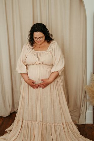 Woman wearing a flowy cream-colored prairie dress with puff sleeves stands against a beige curtain backdrop.