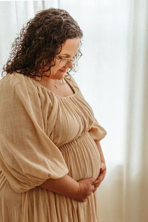 A person in a beige linen dress stands by a window in a series of intimate maternity photos.