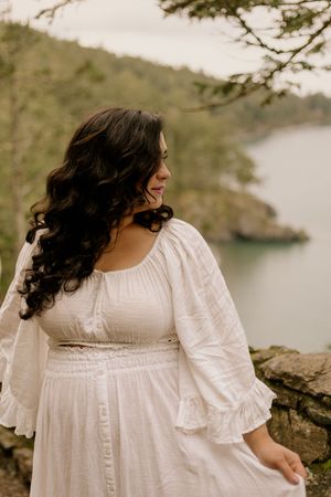 Person in flowing white dress with ruffled sleeves poses on rocky shore near tranquil lake.