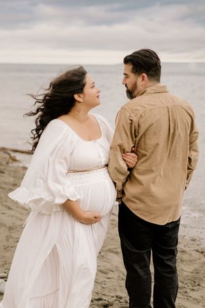 A couple in white dress and brown shirt share intimate moments on a cloudy beach in a romantic photo series.