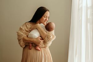 A woman in a beige dress holds a baby near a window with soft natural lighting.