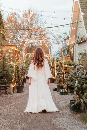 A person in a flowing white wedding dress twirls in a garden decorated with twinkling string lights at dusk.