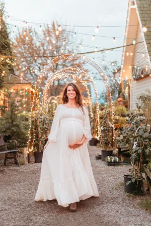 Maternity photo in white dress under twinkling garden lights and floral arches at dusk.