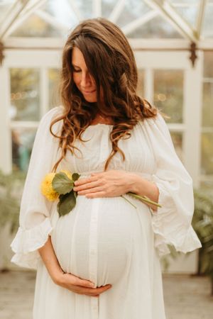 Expectant mother in white flowing dress holds yellow flower in sunlit greenhouse setting.