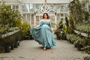 A person in a flowing turquoise dress twirls gracefully in a greenhouse filled with potted plants and natural light.